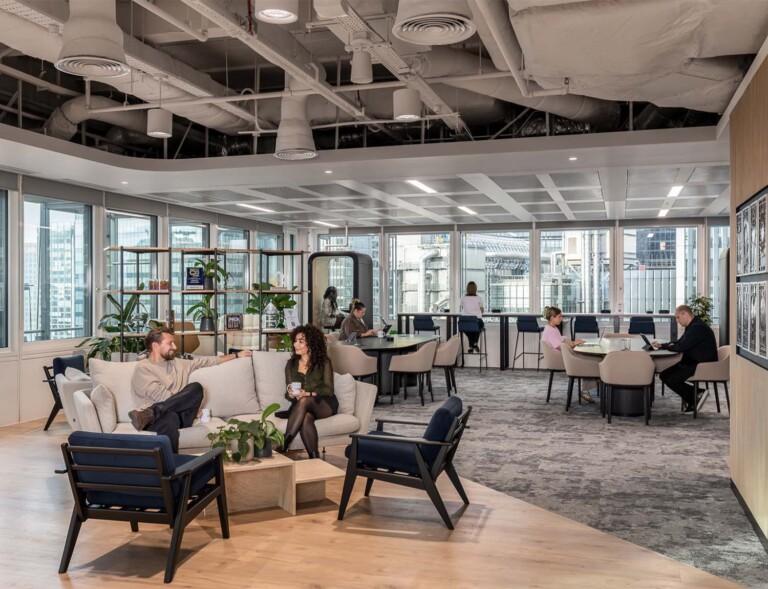 A modern open-plan office lounge with seating areas, plants, and large windows overlooking a city skyline.