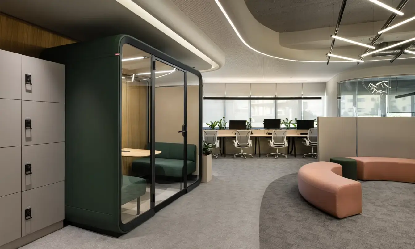 Modern open-plan office with a glass-walled Framery Four meeting pod, lockers on the left, and curved pink seating.