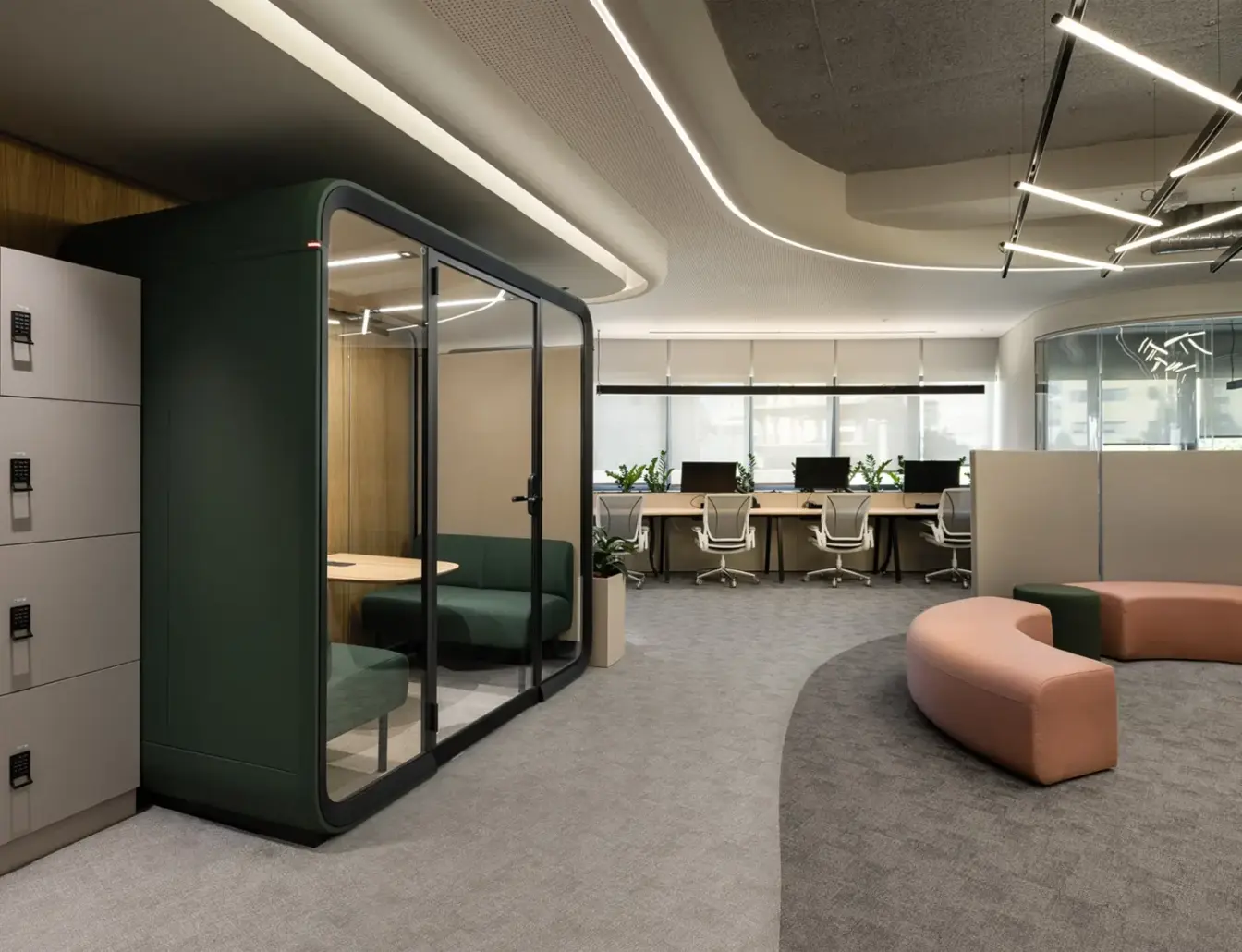 A modern open-plan office with a glass-walled Framery Four meeting pod, lockers, curved pink seating, and desks in the background.