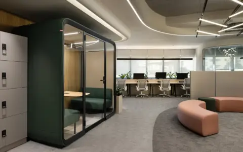 A modern open-plan office with a glass-walled Framery Four meeting pod, lockers, curved pink seating, and desks in the background.