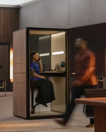 A woman in a blue blouse sits inside a Framery Gradus Focus smart and soundproof focus pod with a laptop, talking on a phone.