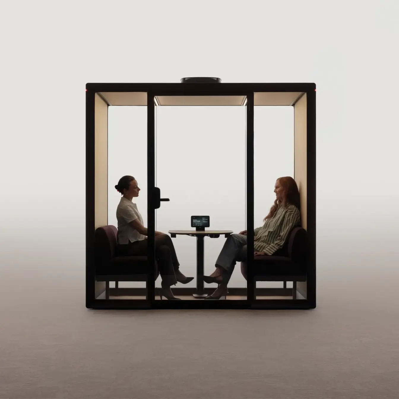 Two people sit facing each other inside a Framery Gradus huddle smart and soundproof huddle pod with a table between them.