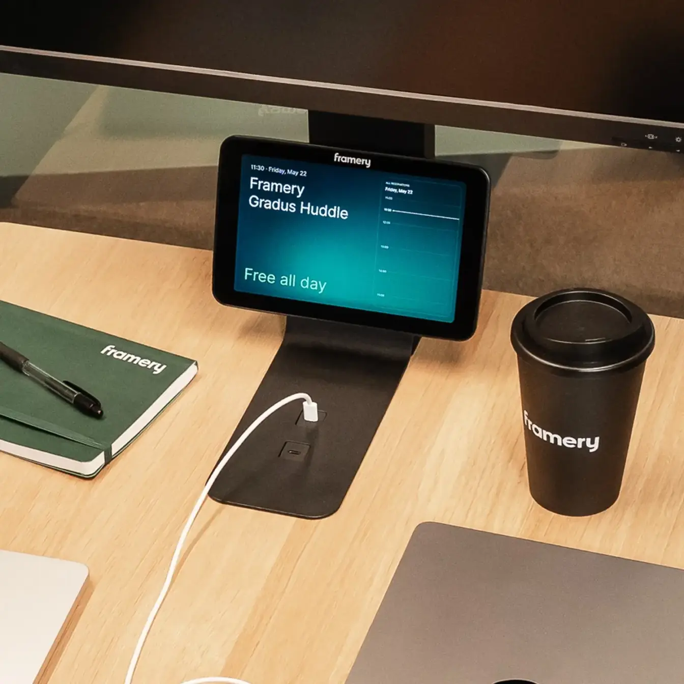 A wooden desk with a small tablet on a stand displaying a blue screen, a black Framery travel mug, a Framery notebook, and a laptop with a charging cable.