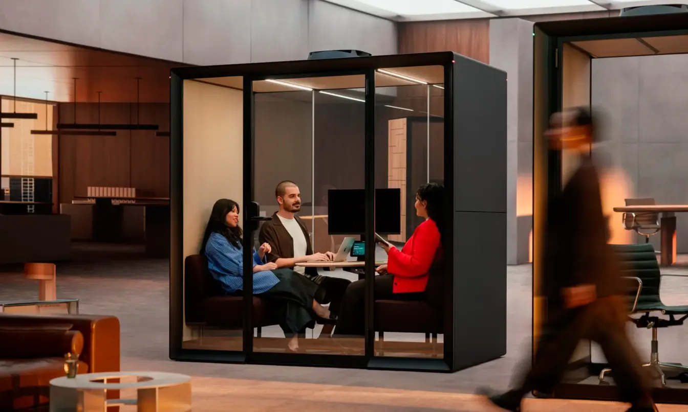 Four people sit around a table inside a glass-walled Framery Gradus Huddle smart and soundproof huddle room in an office, while a person walks by outside.