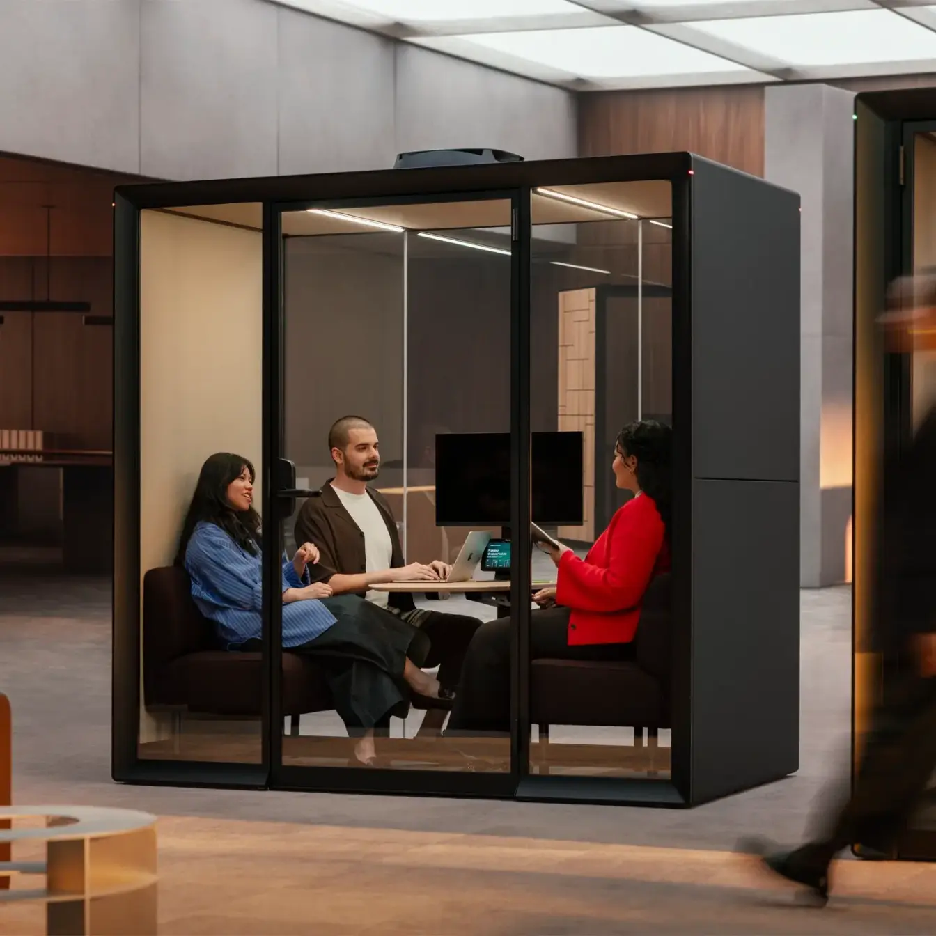 Four coworkers sit around a table inside a glass-walled Framery Gradus Huddle room in a modern office.