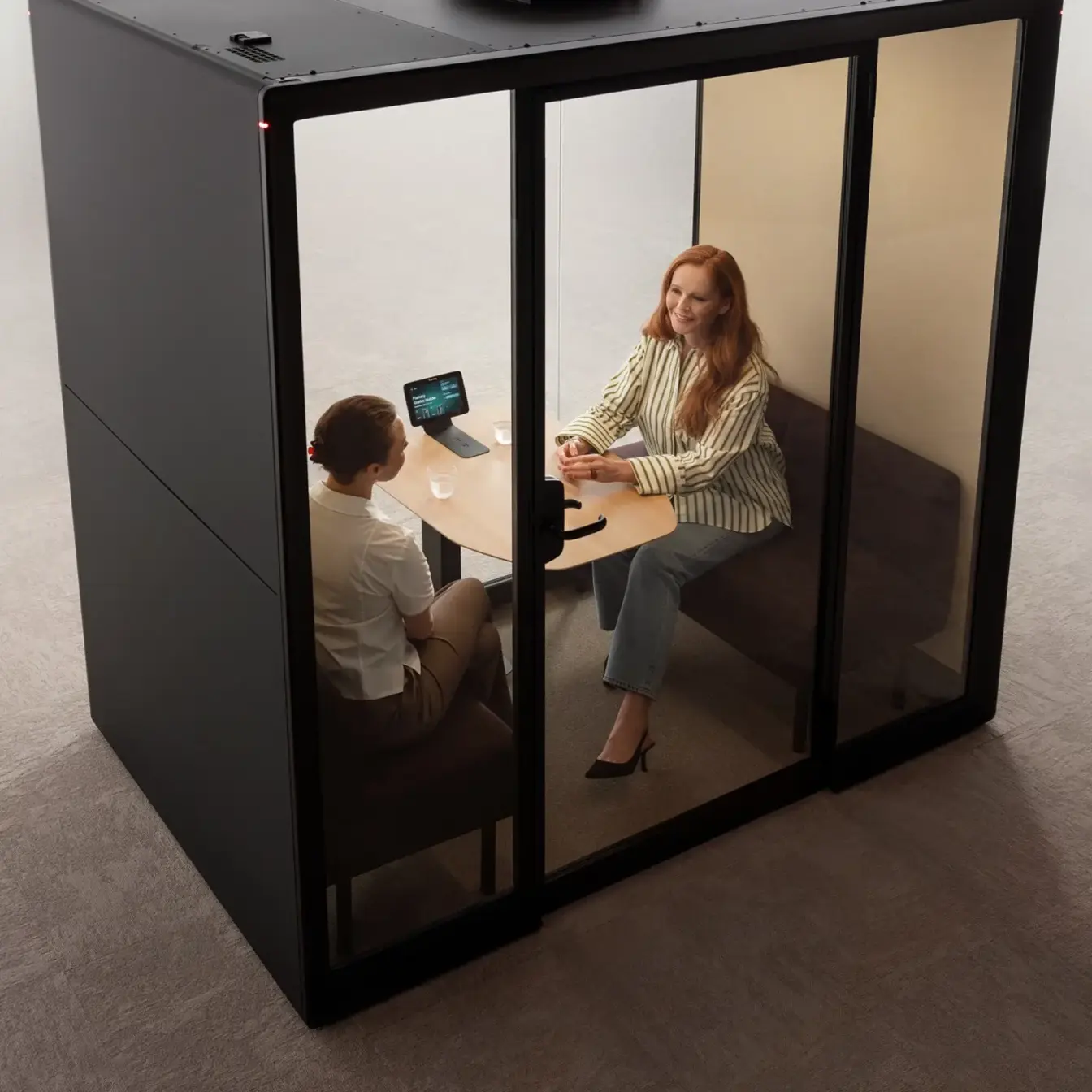 Two people sit across from each other at a small table inside a glass-walled Framery Gradus Huddle smart and soundproof huddle room.