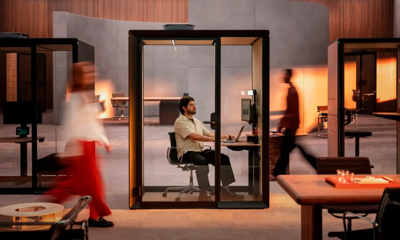 A person working in a Framery Gradus Work. The work pod is located in a busy work environment with people working in an open plan office around the pod.