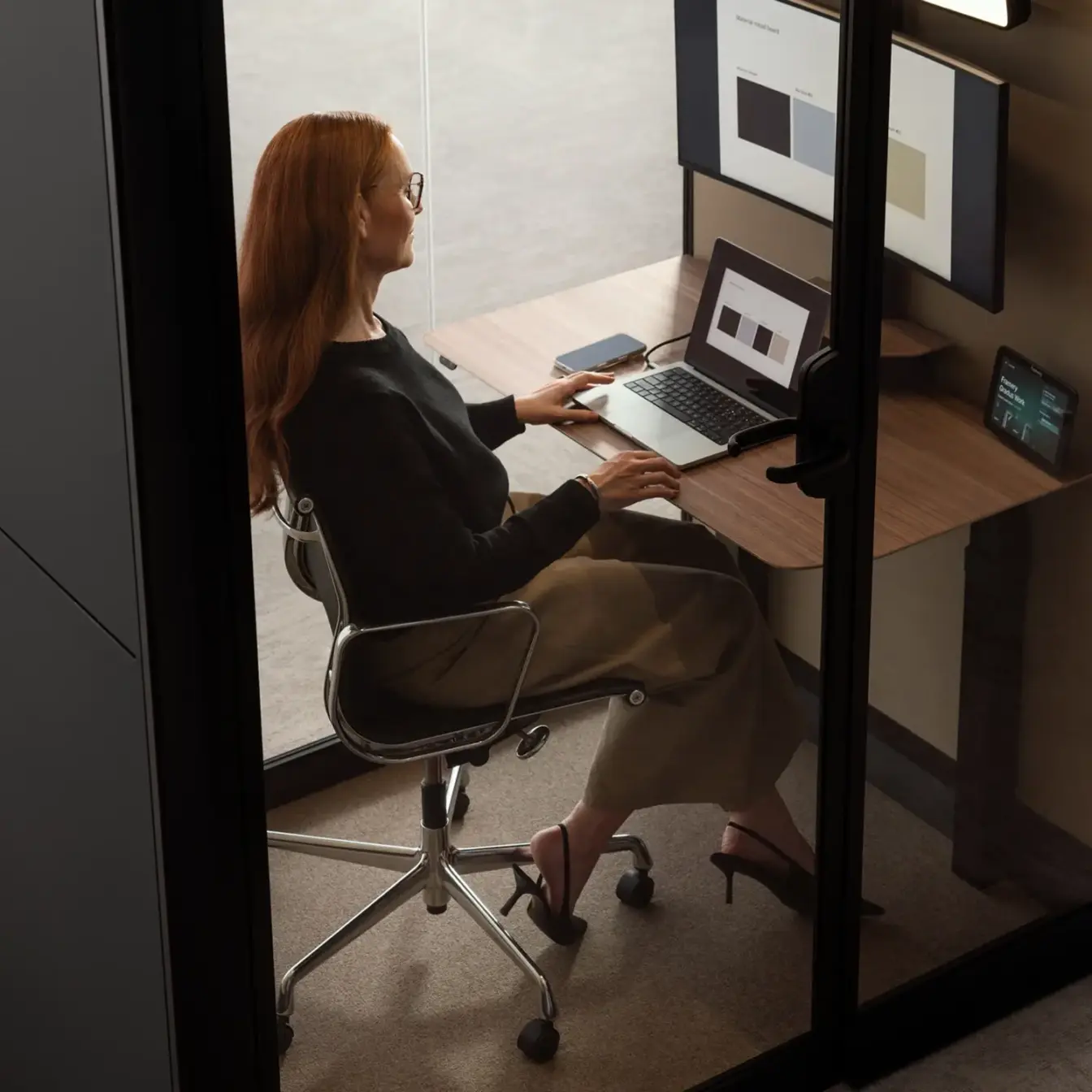 Woman with red hair sits at a desk in a Framery Gradus Work smart work pod, working on a laptop with an external monitor.
