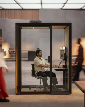 A man sits at a laptop inside a Framery Gradus Work smart and soundproof work pod in a busy workspace.