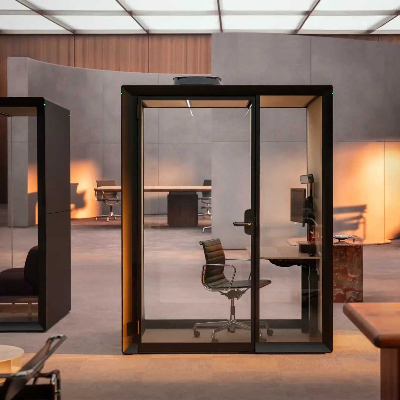 A glass-walled private office booth with a desk and chair inside a modern open workspace.