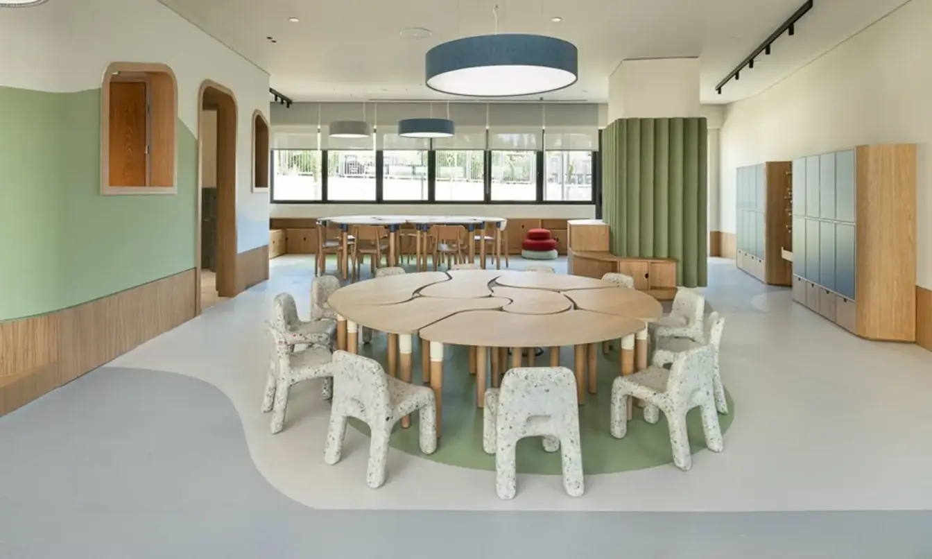 Bright open area meeting space with a large circular table surrounded by white plastic chairs, pastel walls, and long windows along the back wall.