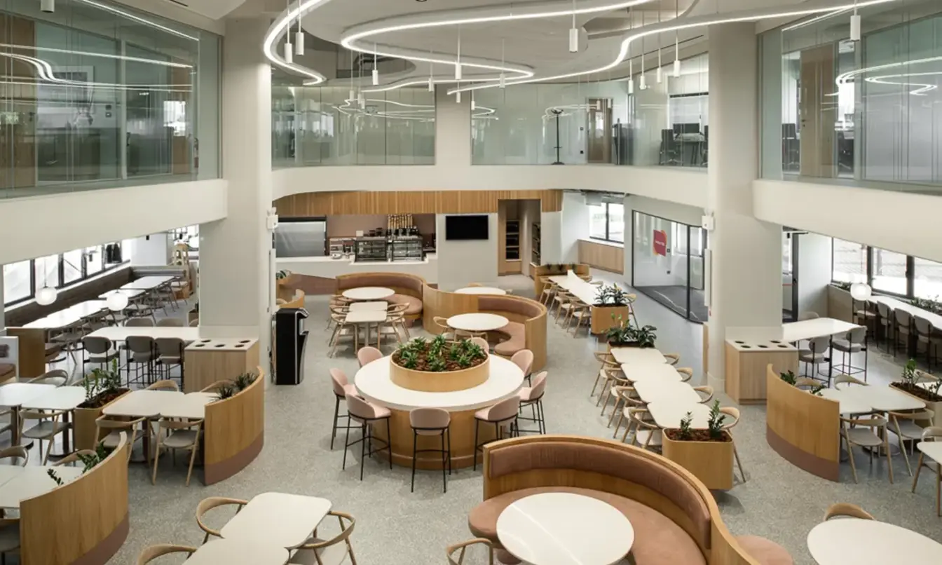 A bright, modern cafeteria interior with circular seating booths around a central plant, long tables along the windows, and an upper mezzanine.