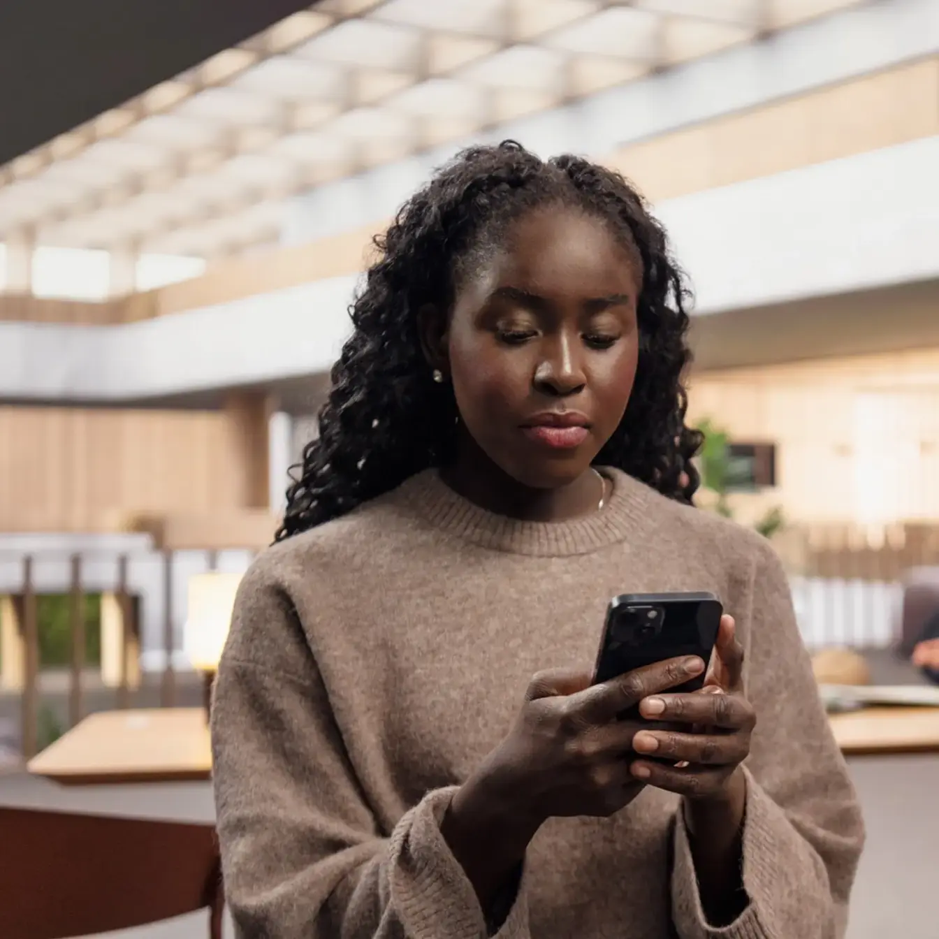 A young woman with curly hair wearing a beige sweater looks at her phone in a bright, modern indoor space.