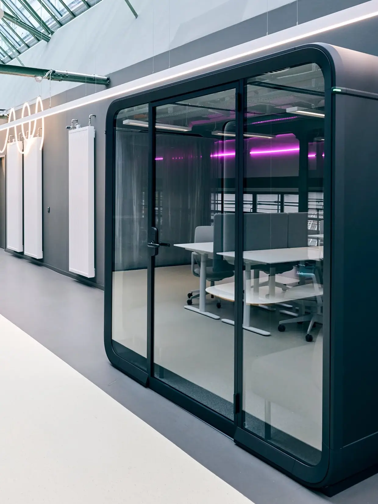 A black framed Framery Four meeting pod set in a light office space with office desks reflecting from its glass wall.
