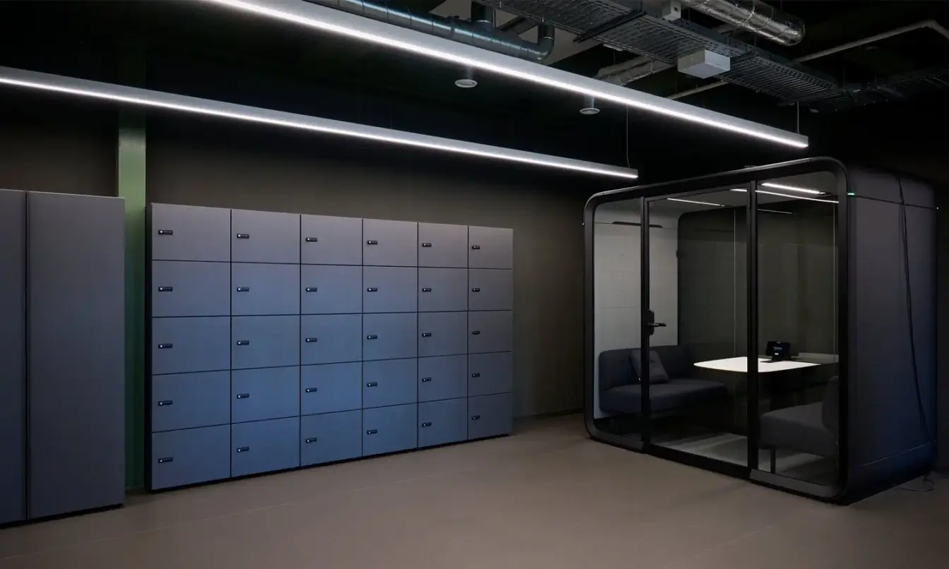 A dark color hallway with a row of dark grey lockers and a Framery Four meeting pod at the back wall.