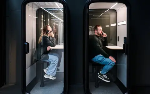 A womand and a man sitting inside two black Framery One Compact phone booths that are lit inside, with a dark wall as background.