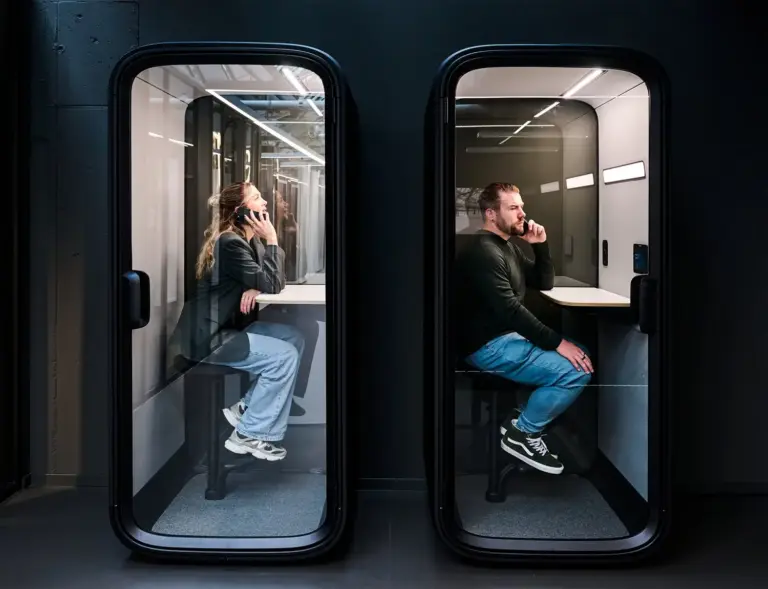 A womand and a man sitting inside two black Framery One Compact phone booths that are lit inside, with a dark wall as background.