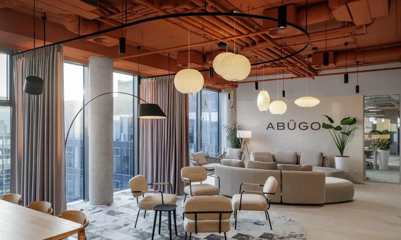 A bright, modern coworking lounge with beige sofas, round chairs, hanging pendant lights, and large windows.