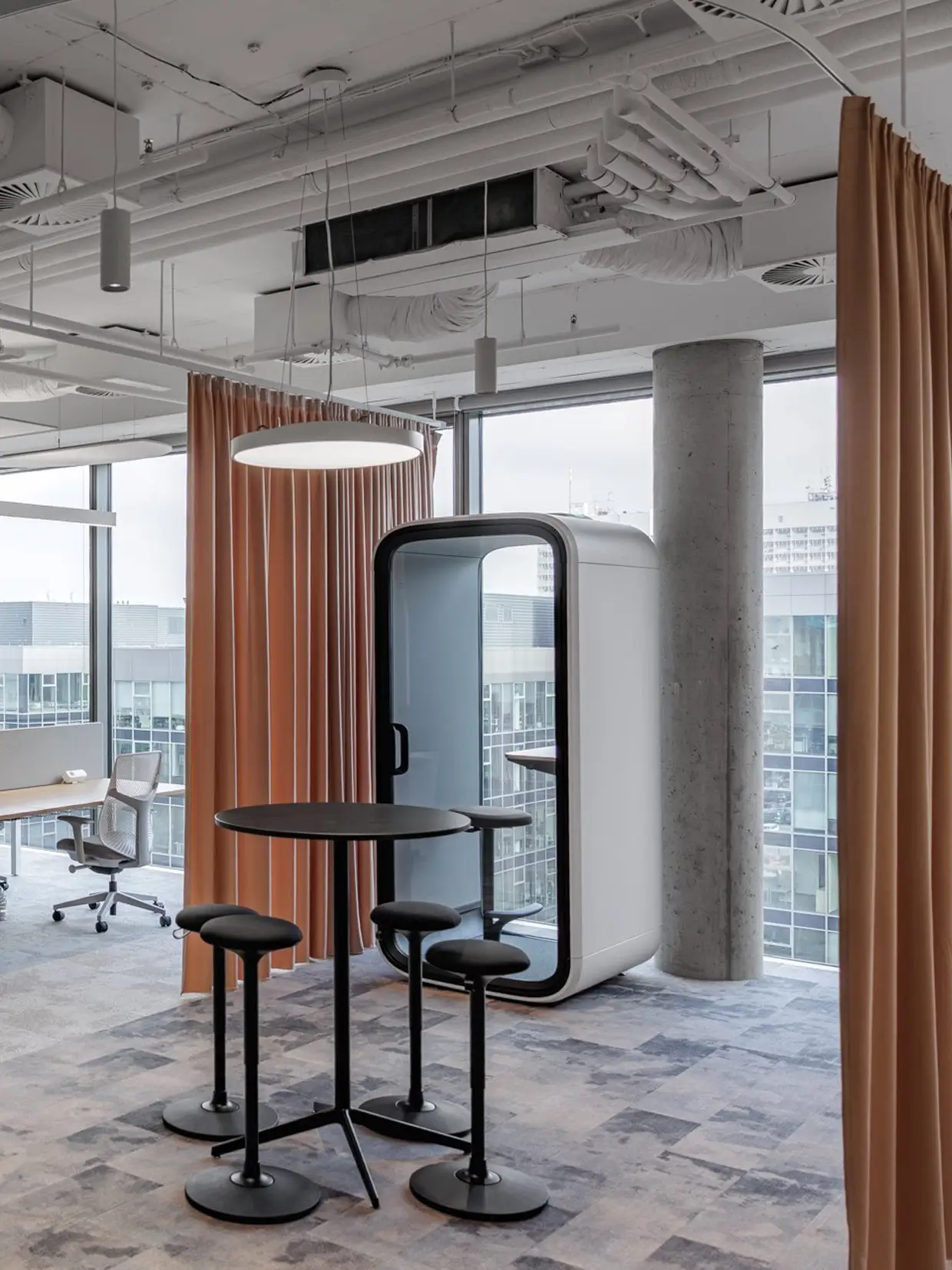 A modern office lounge featuring a white Framery office pod with a round table and two stools beside large windows and orange curtains.