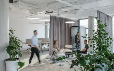 Bright open-plan office with white walls, large green plants, and several people seated around a small table while another person walks by and one person sits inside a white Framery One pod.