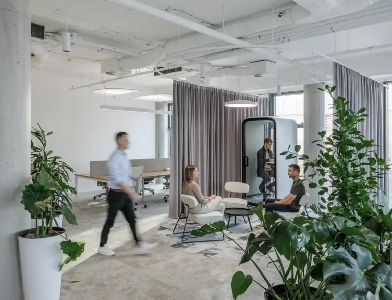 Bright open-plan office with white walls, large green plants, and several people seated around a small table while another person walks by and one person sits inside a white Framery One pod.
