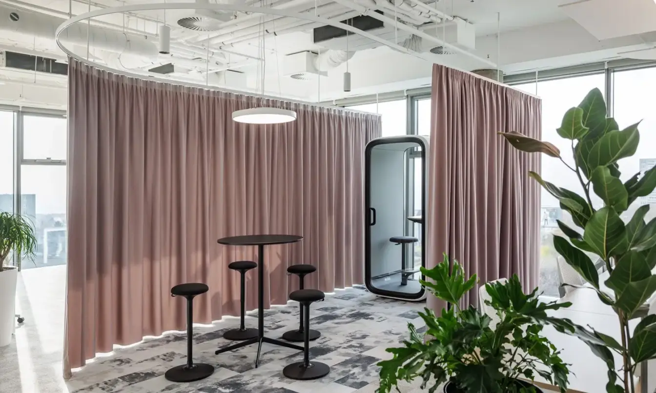 A bright modern office corner with pink partition curtains, black high tables and stools, a white Framery One pod, and several potted plants.