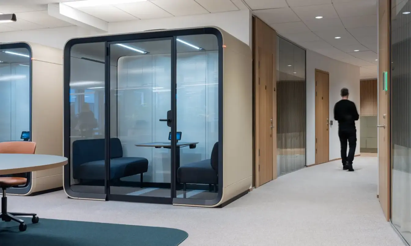 An curved office hallway with a man walking away and a sand colored Framery Four meeting pod around the corner.