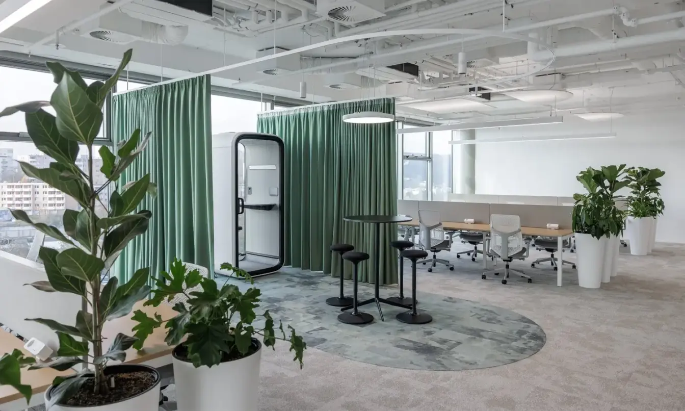 Open-plan office with greenery, a green curtain partition around a standing work station on a circular rug and a Framery phone booth, and desks and chairs in the background.