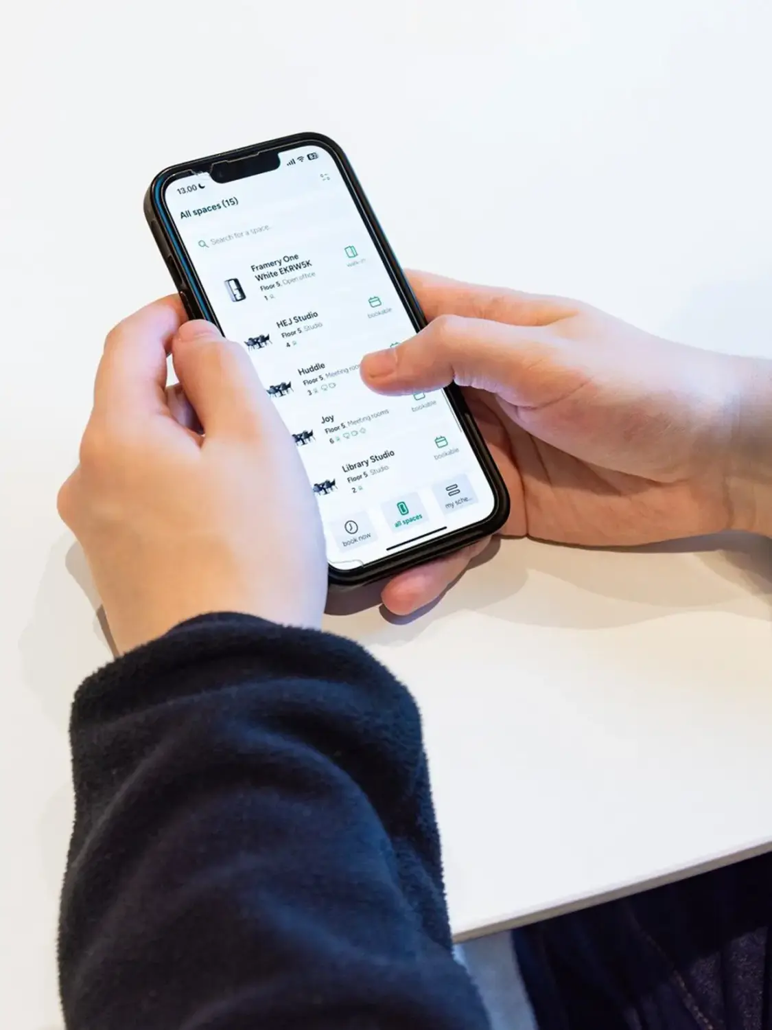 Two hands hold a smartphone displaying a list of available meeting rooms in Framery App on a white surface.