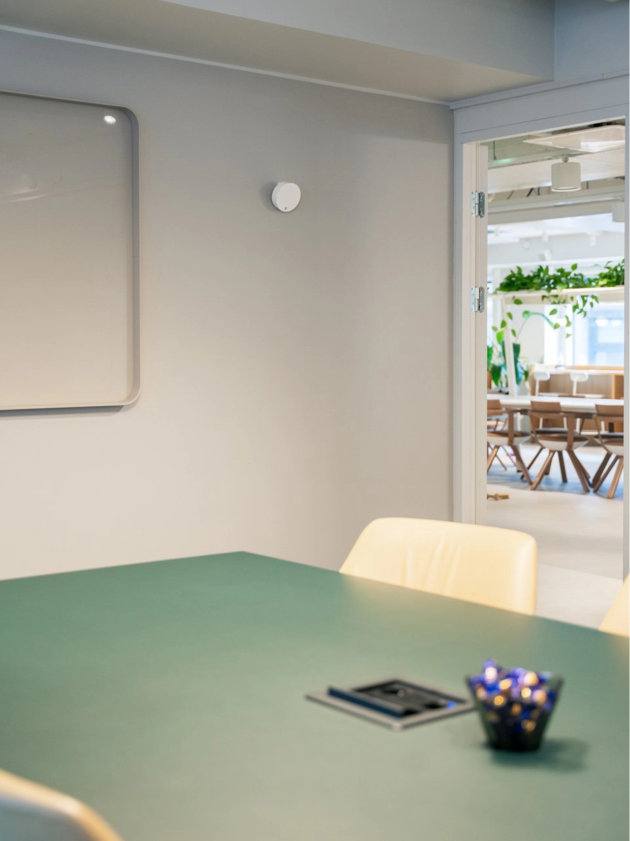 Bright meeting room with a green table in the foreground, a beige chair, a white Framery Room Sensor on the wall and an open doorway to a sunny seating area with plants.