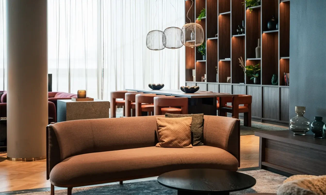 Modern hotel living area with a curved brown sofa, round coffee table, and a shelving wall with plants in the background.