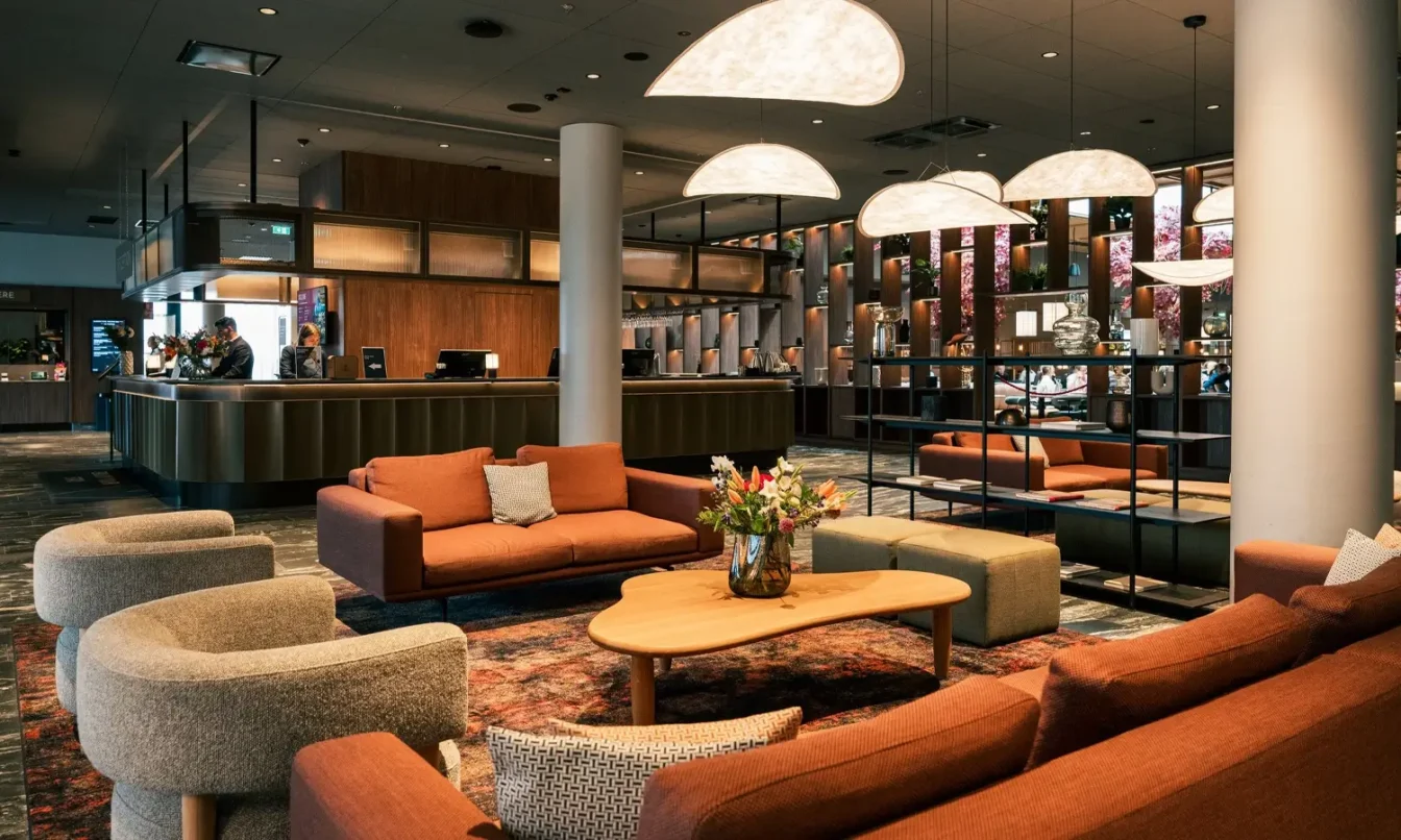 A modern hotel lobby lounge with orange and beige seating around a round coffee table and a flower arrangement, with a reception area in the background.