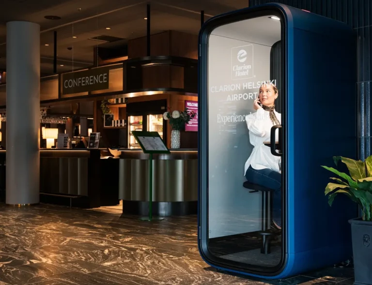 A person sits inside a blue Framery One Compact phone booth in a modern lobby near a conference desk.
