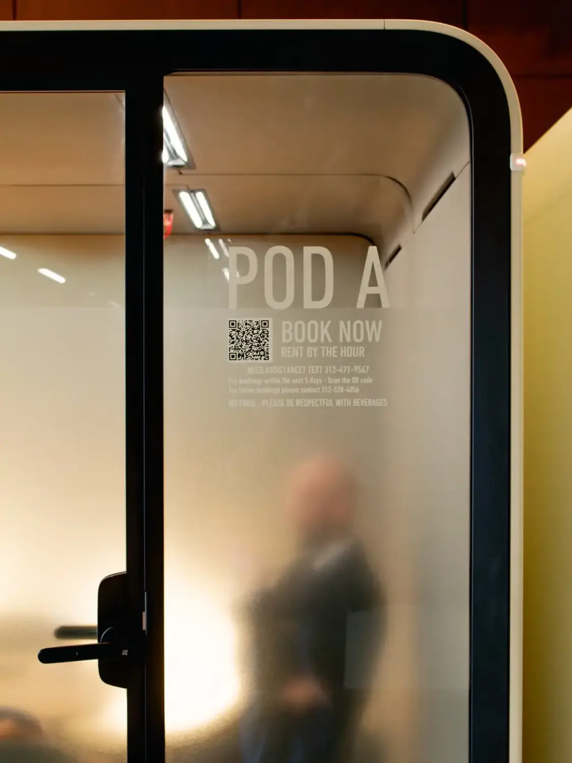 Framery Six meeting room's glass door labeled POD A with a QR code and “BOOK NOW” text, showing a blurred figure inside.