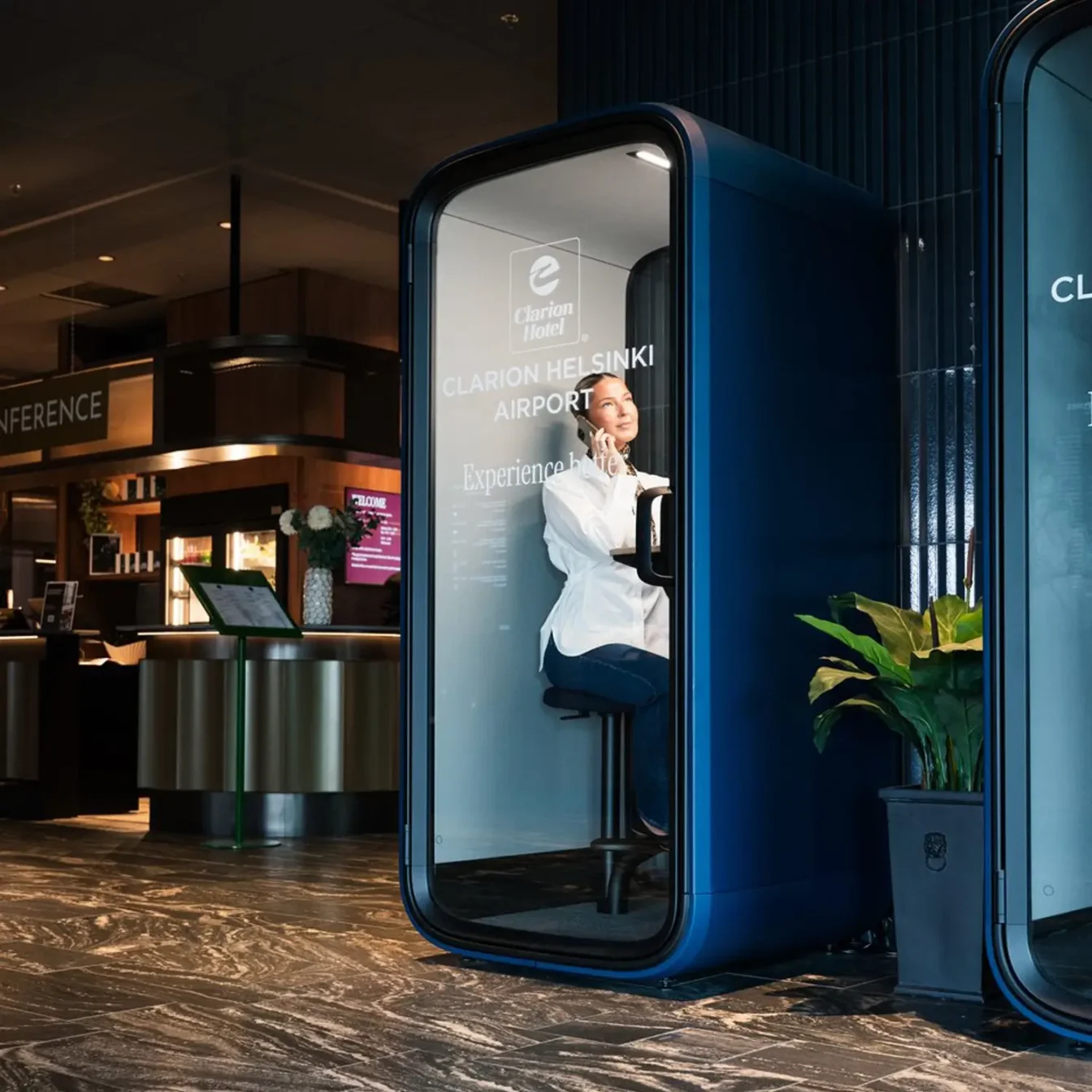 A person sits inside a blue Framery Subscribed phone booth in a modern airport lobby, using a mobile phone.