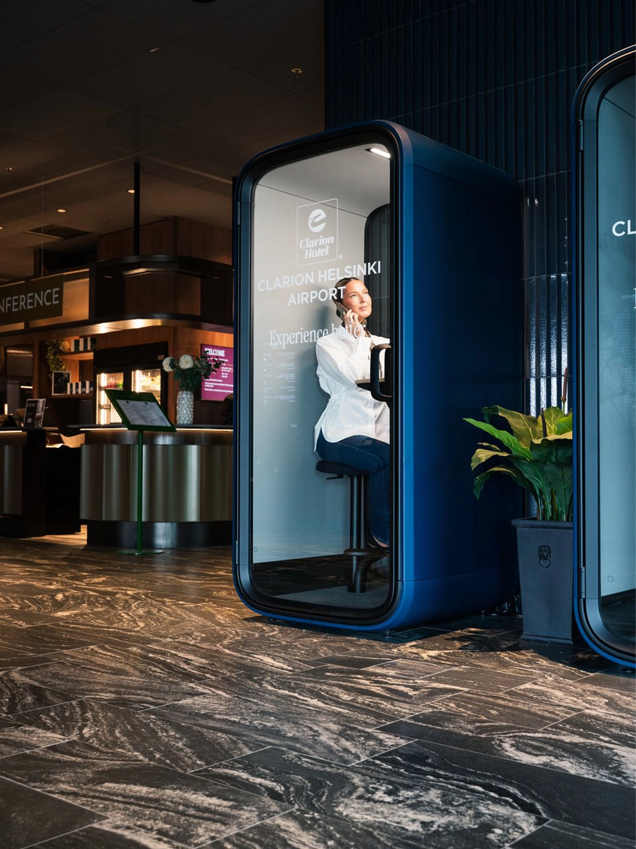 A person sits inside a blue, Framery Subscriped renal phone booth in a modern lobby with a plant and marble floor.
