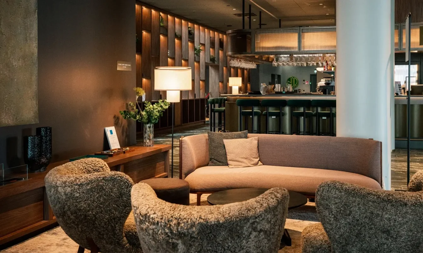 A modern hotel lounge with a curved beige sofa, plush gray chairs, a wooden coffee table, and a bar in the background.