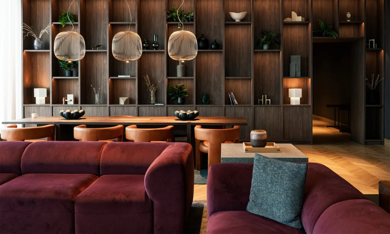 Cozy contemporary lounge featuring a dark wood wall of open shelves, a long wooden table, and burgundy sofas with cushions under round pendant lights.