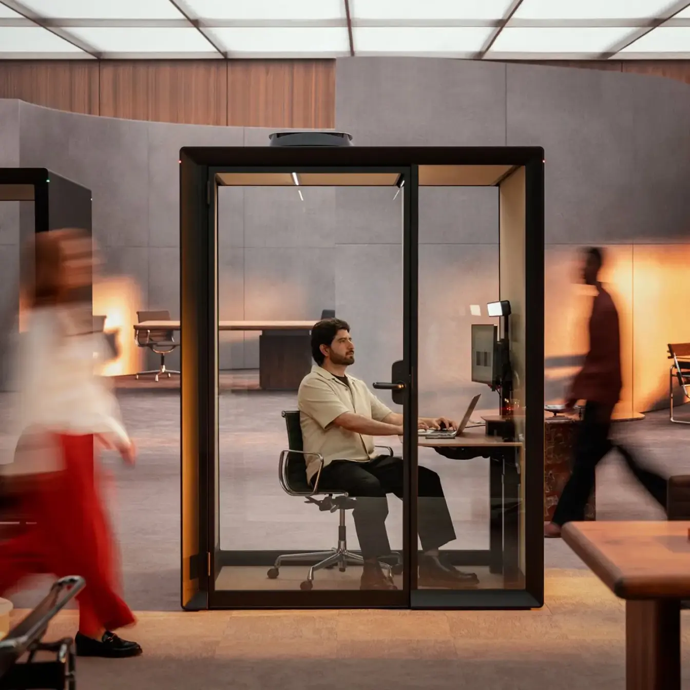 A man sits at a desk inside a glass-walled Framery Gradus Work work pod in a busy open workspace, with blurred people walking by.