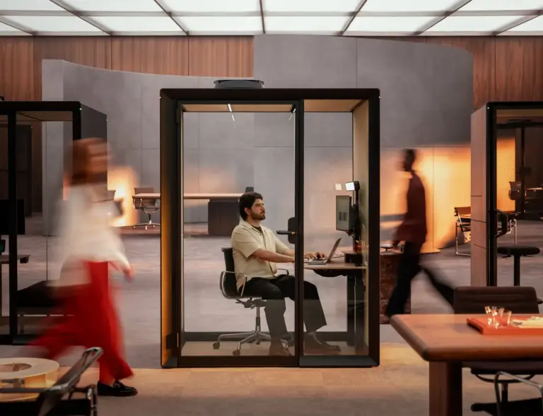 A man sits at a desk inside a glass-walled Framery Gradus Work work pod, while blurred coworkers walk by in a modern open-plan workspace.