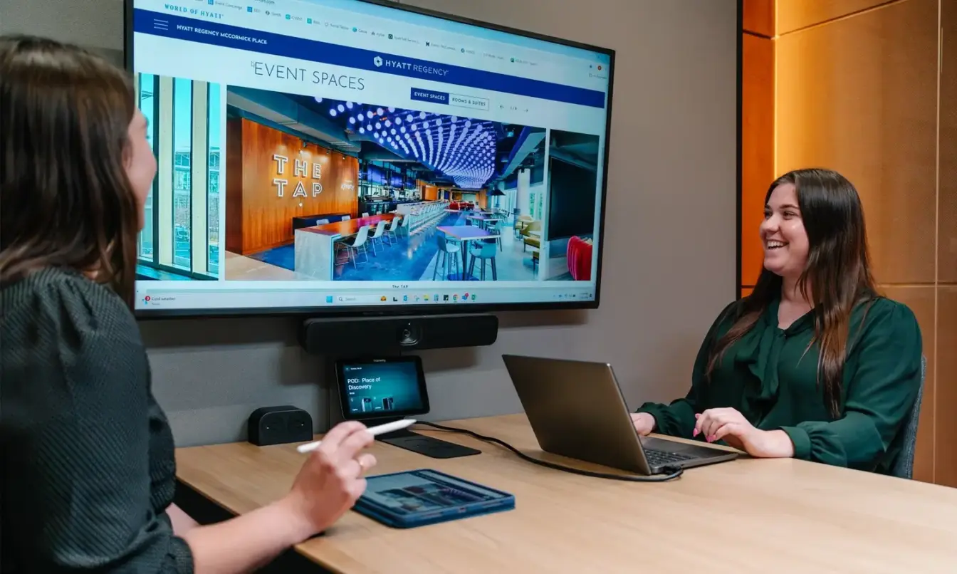Two women in a Framery Six soundproof meeting room sit at a conference table, one with a laptop, discussing a presentation on a wall-mounted screen.