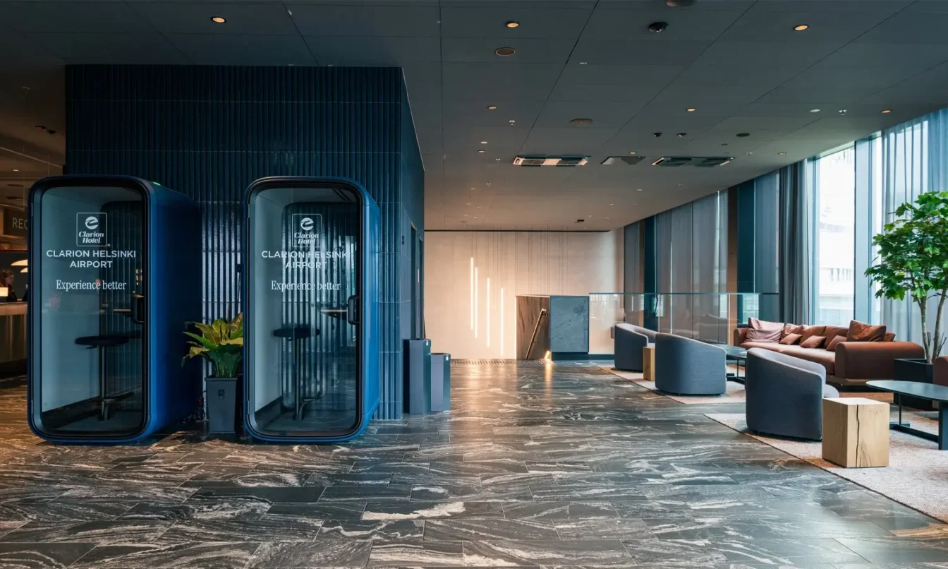 A modern hotel lobby with blue Framery One Compact phone booths on the left, a marble floor, and a seating area of sofas along large windows on the right.