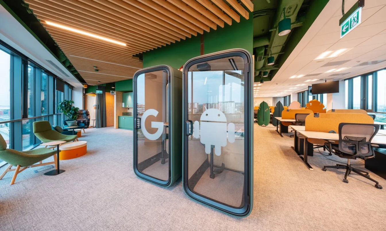 Open-plan office space with two Framery One Compact smart pods displaying the Android logo and Google “G,” surrounded by modern seating, plants, and large windows.