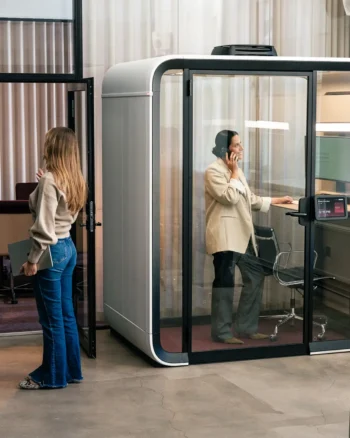 A woman inside a Framery One Work smart workstation pod in an office talks on her phone, while another woman stands outside near the door.