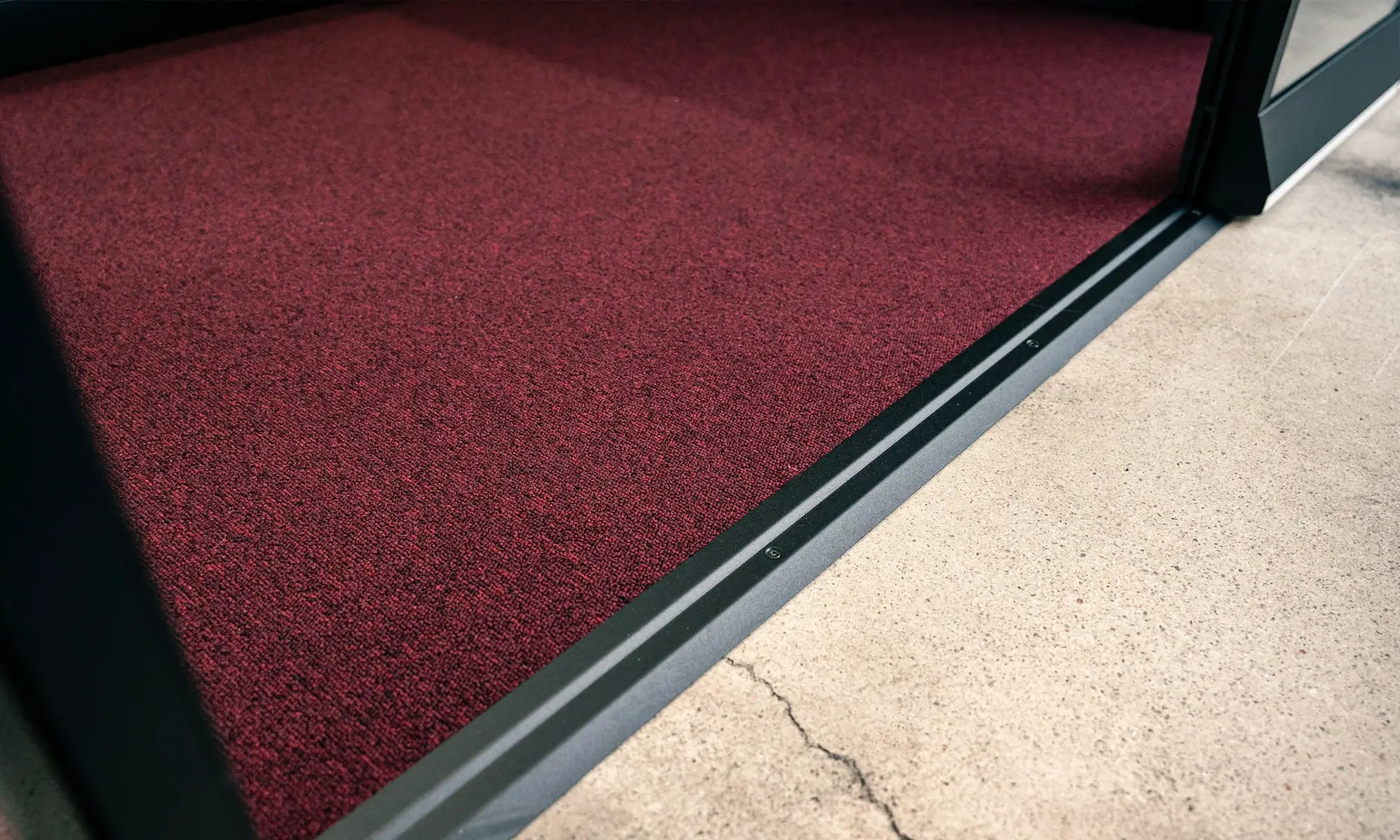 A red carpet mat inside an open doorway of a Framery One Work workstation pod with a metal threshold and concrete floor outside.