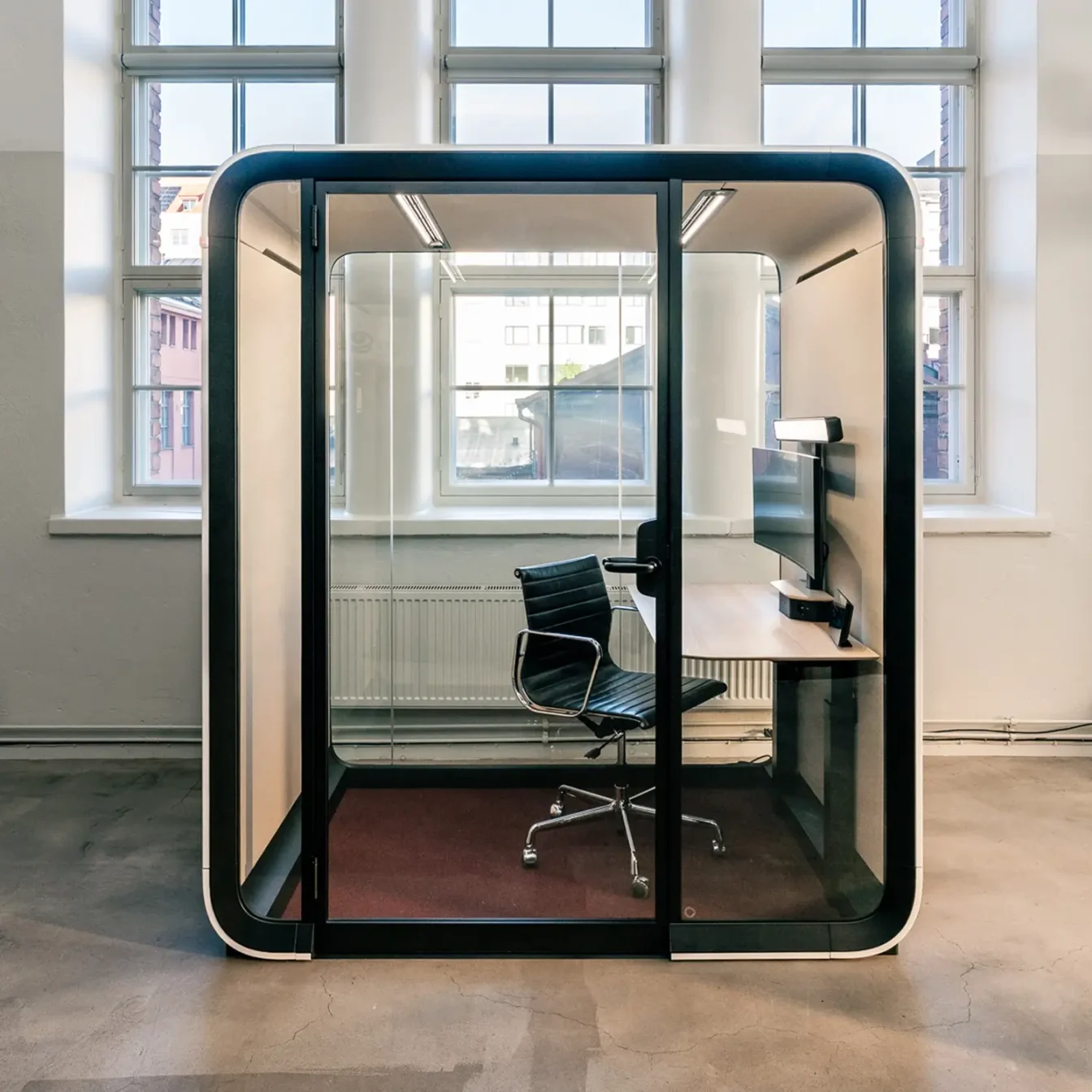 A glass-walled Framery One Work workstation pod with a desk, chair, and computer monitor inside a bright, spacious room.