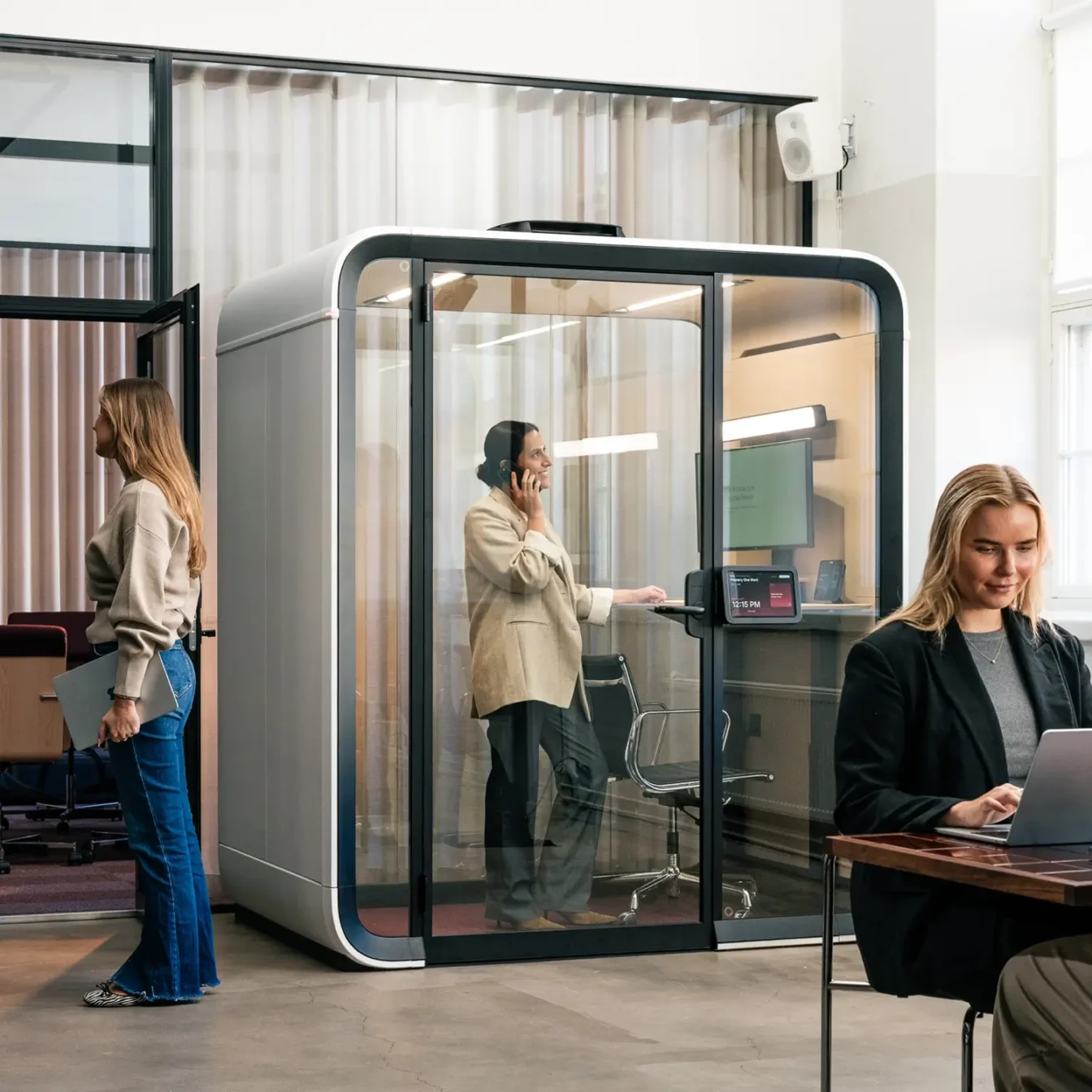 Modern office with a glass-walled Framery One Work workstation pod; a woman sits at a laptop in the foreground, while another person stands inside the pod and a third person walks by.