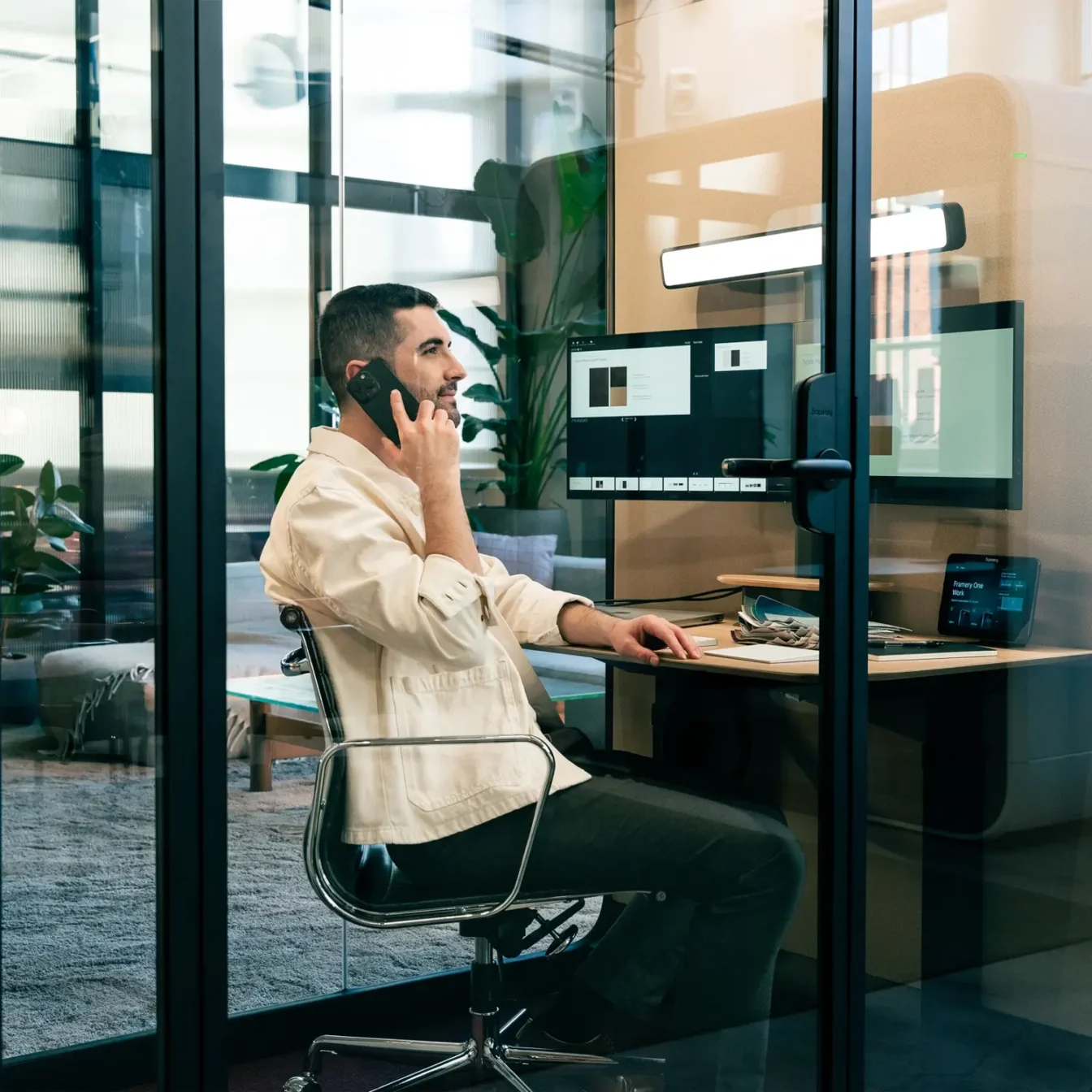 A man sits at a desk in a glass-walled Framery One Work workstation pod, talking on the phone while working at a computer.