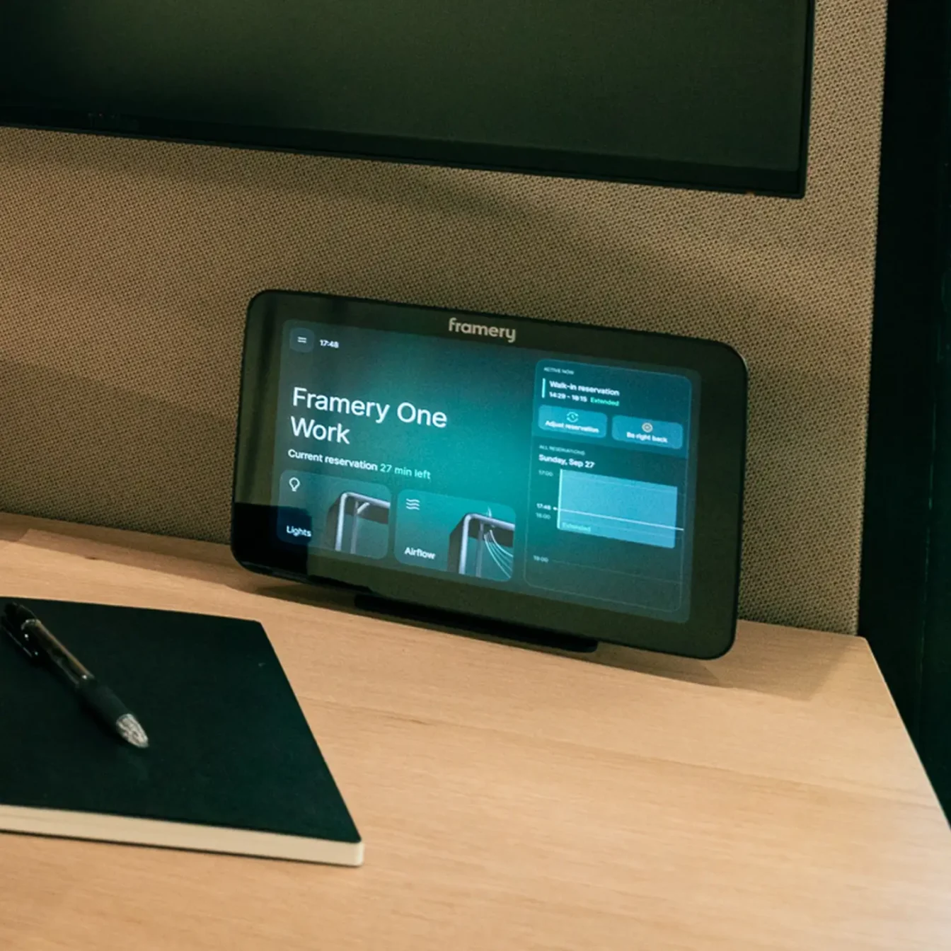 A small display on a wooden desk showing the Framery One Work workstation pod interface, with a notebook and pen nearby.