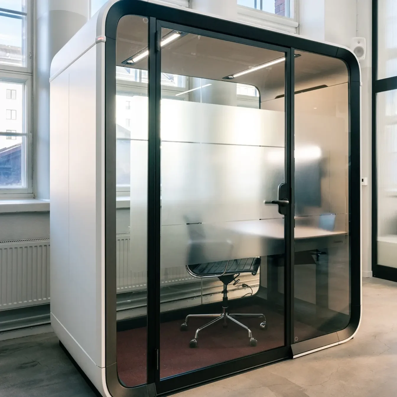 Glass-walled private Framery One Work workstation pod with a desk and swivel chair inside a bright workspace.